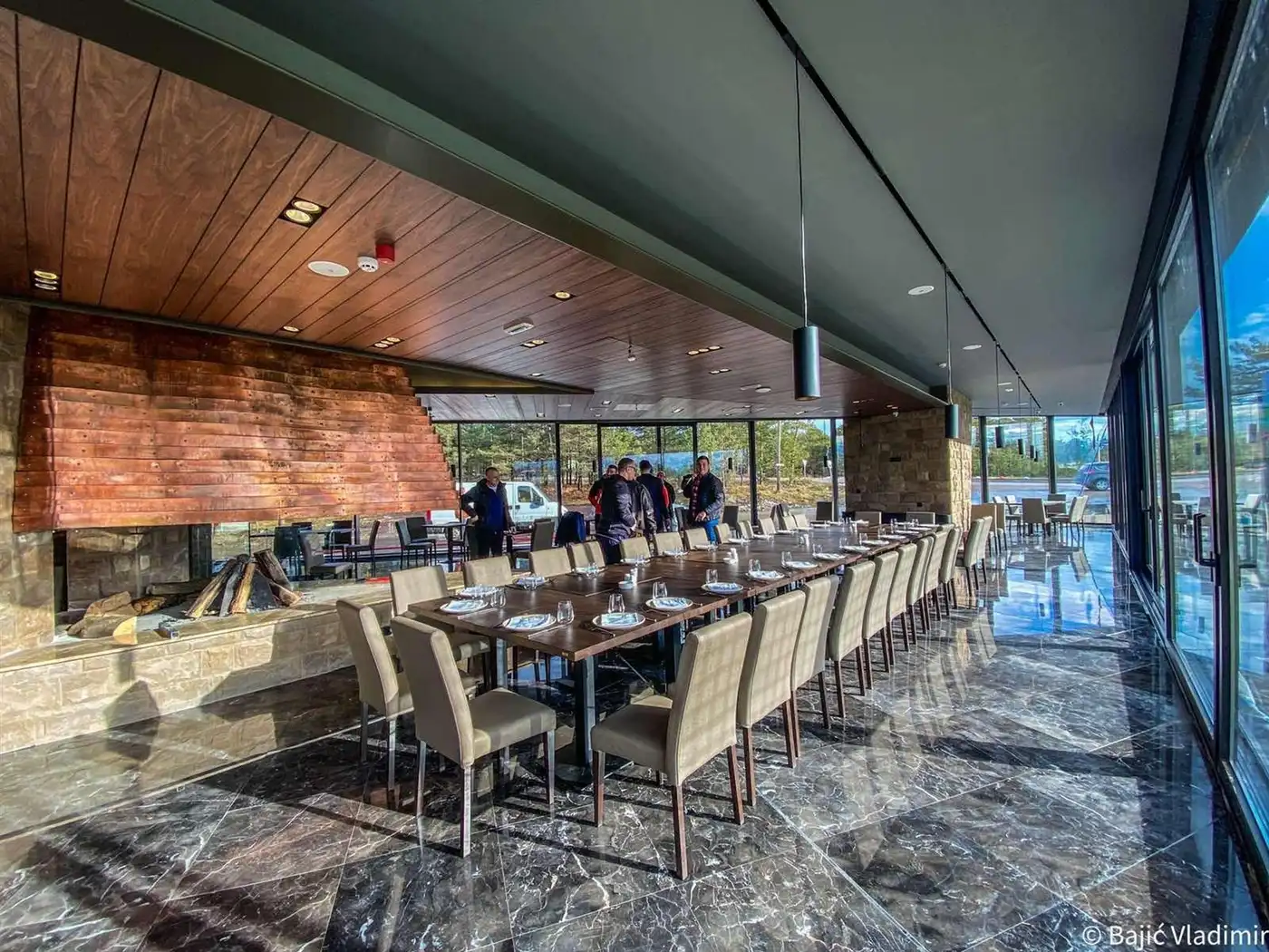 A restaurant room with a long wooden table, a stone fireplace and a glass wall facing the mountain